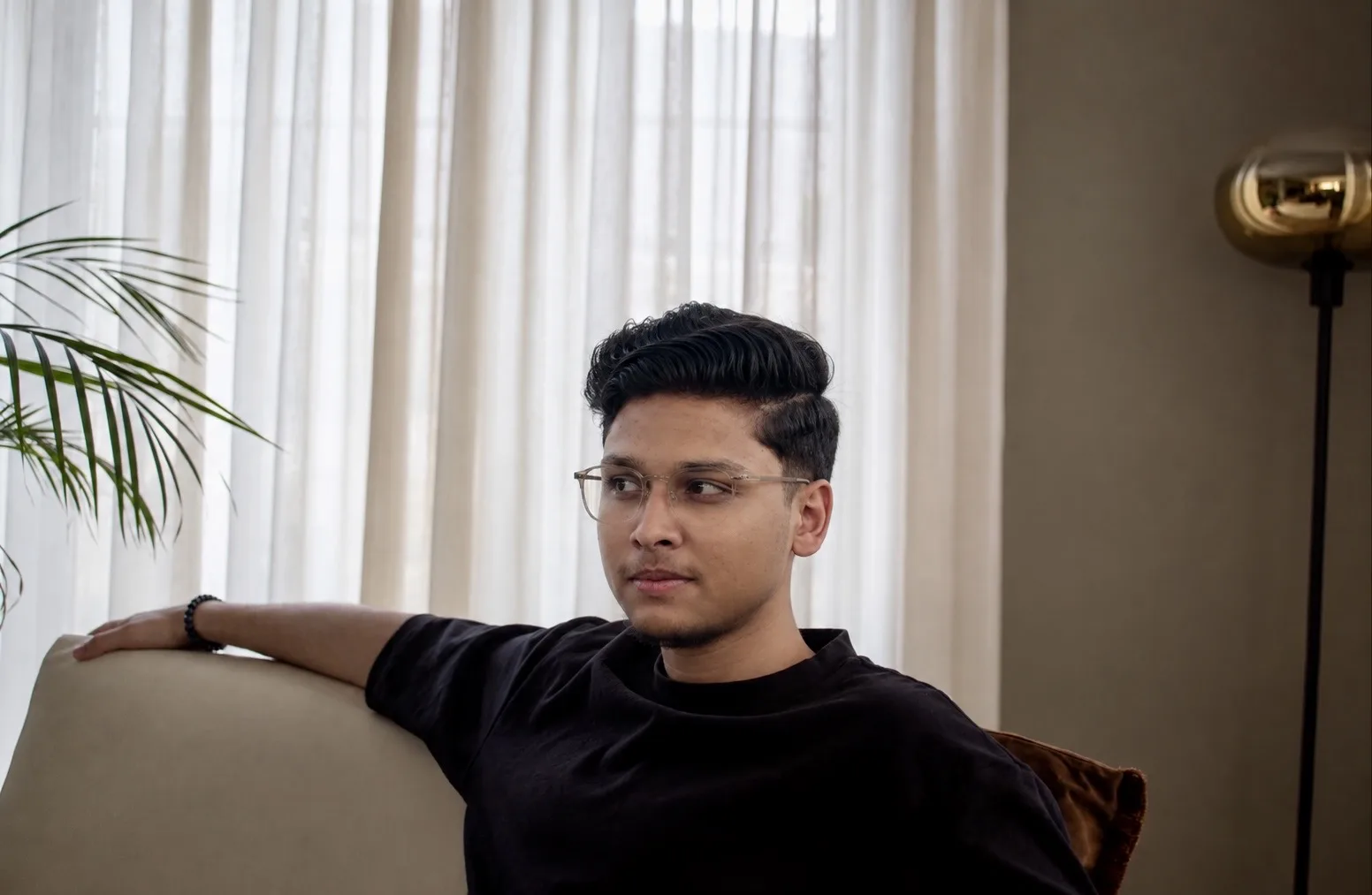 M.Maaz sitting on a couch by sheer curtains indoors.