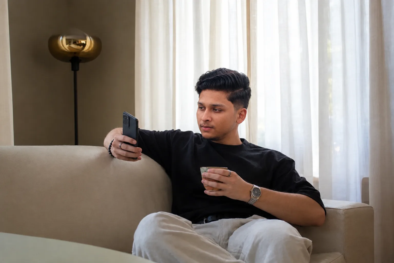 M.Maaz sitting on a couch, looking at his phone and holding a cup.